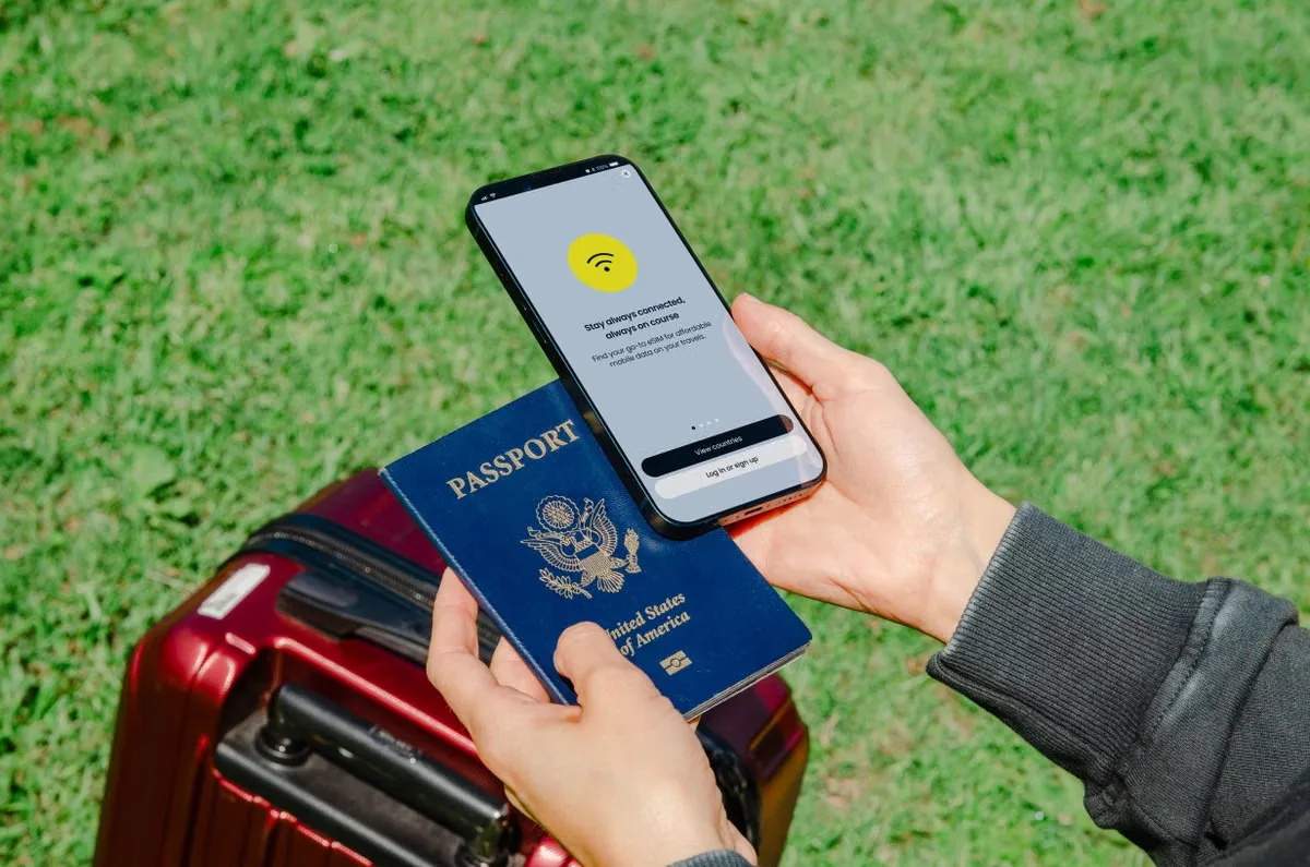 A traveller holding a smartphone showing a stay-connected message, with a passport and suitcase