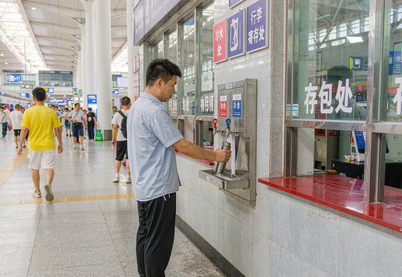 A traveler refilling a reusable bottle at a free hot and cold water dispenser in a Chinese train station